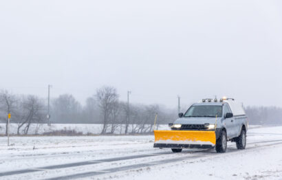 A utility pickup with a yellow blade drives along a snow covered road in open countryside under grey haze. The mood speaks of readiness and safer routes during cold weather.