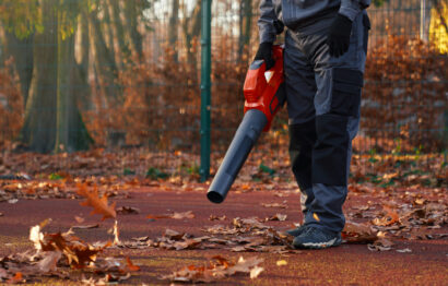 Unrecognizable maintenance man wearing blue uniform and gloves, working with portable leaf blower in morning. Cropped view of male worker holding, using leaf blower at park area. Concept of work.