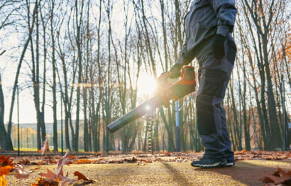 Unrecognizable maintenance worker taking care of court during autumn. Low angle view of male worker in overalls and gloves removing dry leaves with leaf blower against sunlight. Fall, work concept.